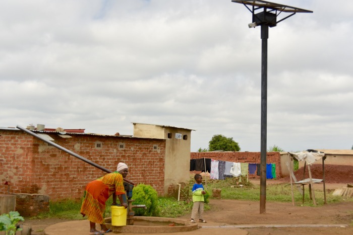 Several finished homes around a nearly complete streetlight installation that also illuminates a drinking-water well, enabling nighttime access to water.
